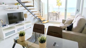 Una sala moderna con televisor de pantalla plana, mesa de vidrio, sofá y una escalera que lleva a un balcón con vista al mar.