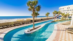 A pool area with a curved design, palm trees, and a beach view. - The Oasis 1506 | Spacious, Lazy River, Poolside Grill, New Splash Pad, Pool with Slide! (Orange Beach)
