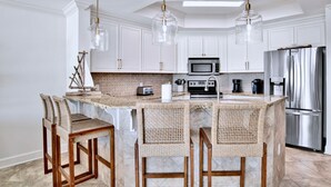 A modern kitchen with white cabinets, a central island with bar stools, stainless steel appliances, and pendant lighting. - The Oasis 1506 | Spacious, Lazy River, Poolside Grill, New Splash Pad, Pool with Slide! (Orange Beach)