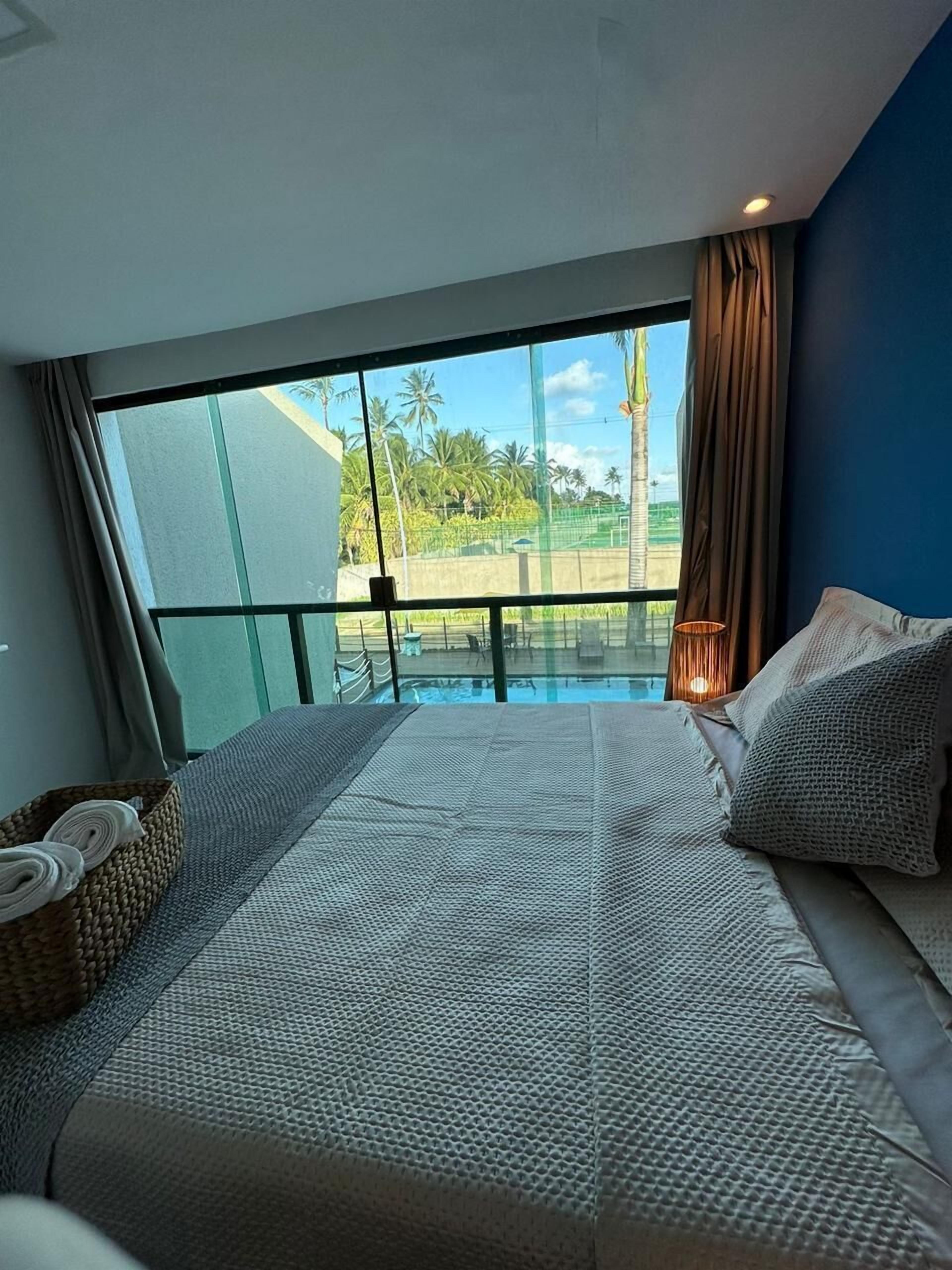 A bedroom with a large bed, a view of a pool and palm trees, and a balcony with a railing.