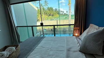 A bedroom with a large bed, a view of a pool and palm trees, and a balcony with a railing.