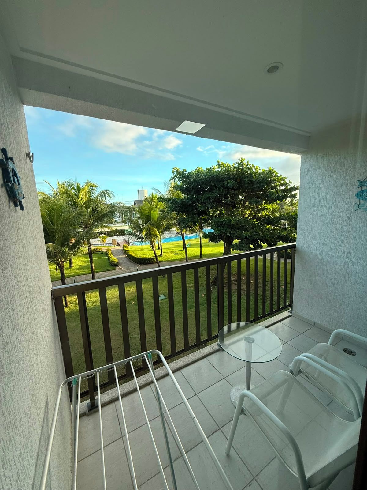 A balcony with a glass table and chairs, a metal railing, and a view of a green lawn with trees and a building in the distance.