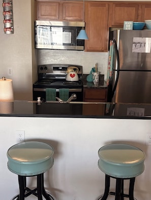 A kitchen with a stainless steel refrigerator, a microwave, and a stove. There are two stools with teal seats and black legs.