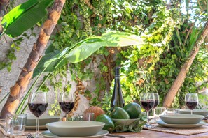 A dining table set outdoors with plates, glasses, and a bottle, surrounded by greenery. - Seabreeze Boho Stay - Apartment with sea view (Letojanni)