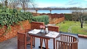 En terrasse med et bord og stole, en murstensmur med grøn bevoksning og udsigt til et sø.
