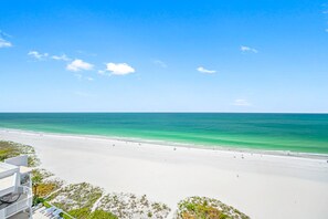 A beach view with a clear sky, sandy shore, and a building with a balcony overlooking the sea.