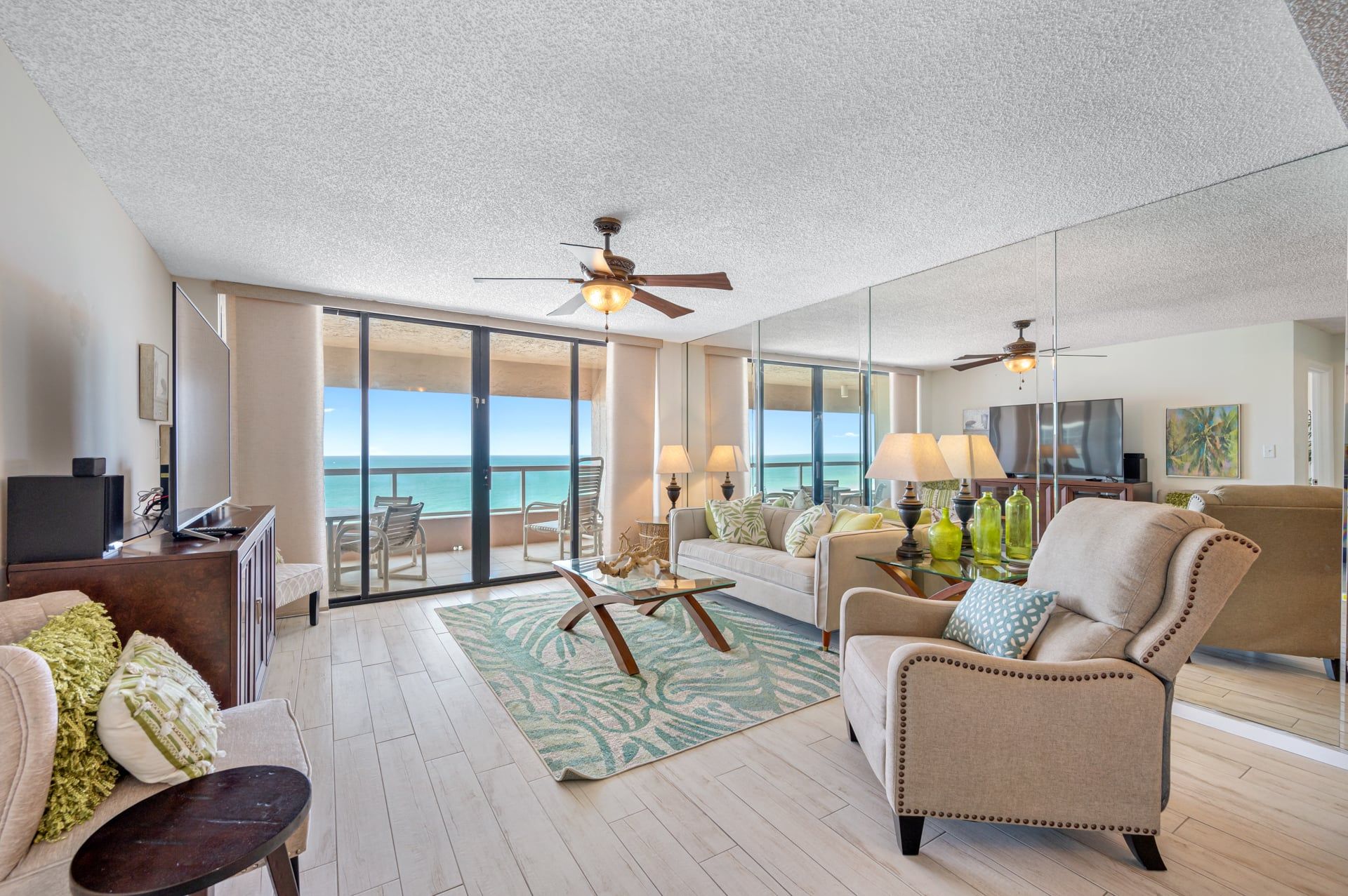 A spacious living room with a large window overlooking the ocean, a ceiling fan, and a flat-screen TV.