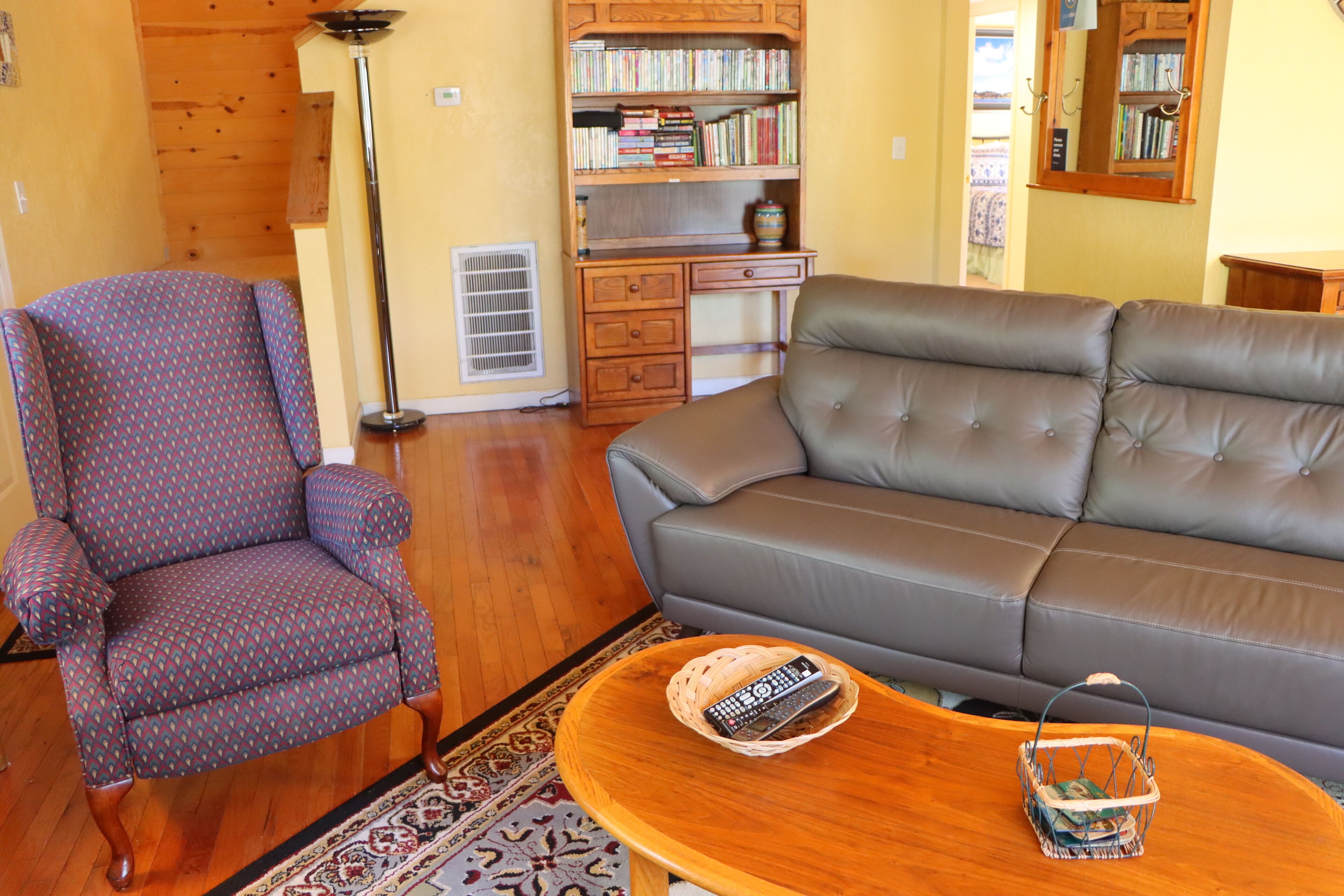A living room with a patterned armchair, a leather sofa, a wooden coffee table with a remote control, and a bookshelf filled with books.