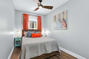 A bedroom with a bed, a ceiling fan, a window with curtains, a nightstand with a lamp, and a wall painting.