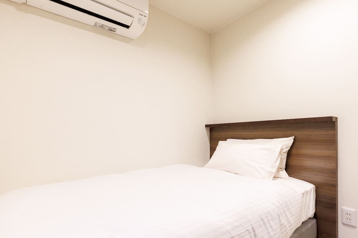 A neatly made bed with a wooden headboard in a room with white walls and a ceiling-mounted air conditioner.