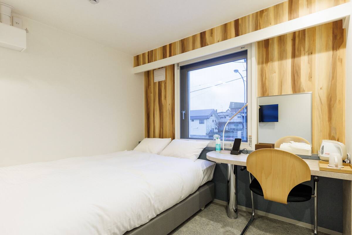 A hotel room with a bed, a desk with a chair, a window, and a view of buildings outside.