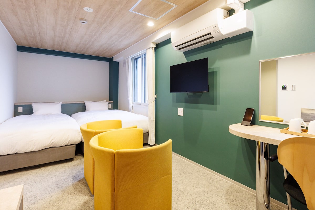 A hotel room with two beds, a desk, a chair, a TV, and an air conditioning unit.