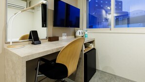 Une chambre d’hôtel équipée d’un bureau, d’une chaise, d’un téléviseur à écran plat, d’un miroir, d’une fenêtre donnant sur des immeubles et d’un petit placard.