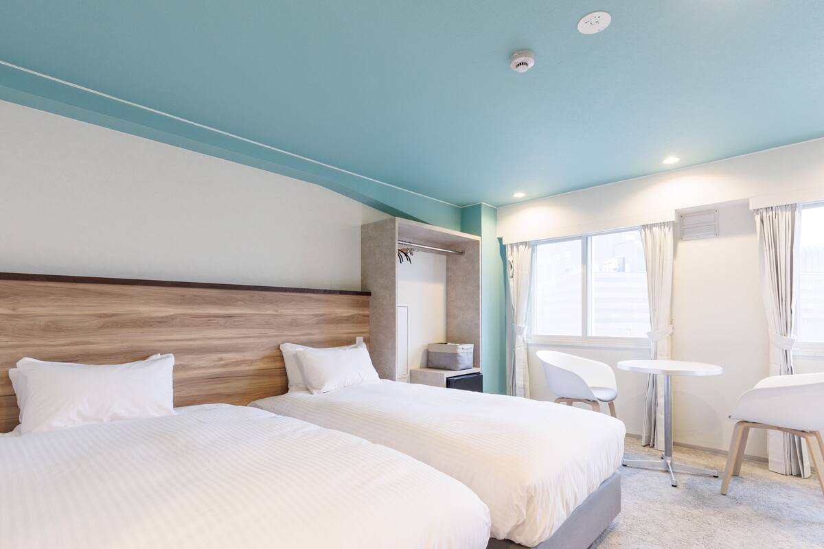 A hotel room with two beds, a small table, a chair, a window with curtains, and a ceiling-mounted smoke detector.