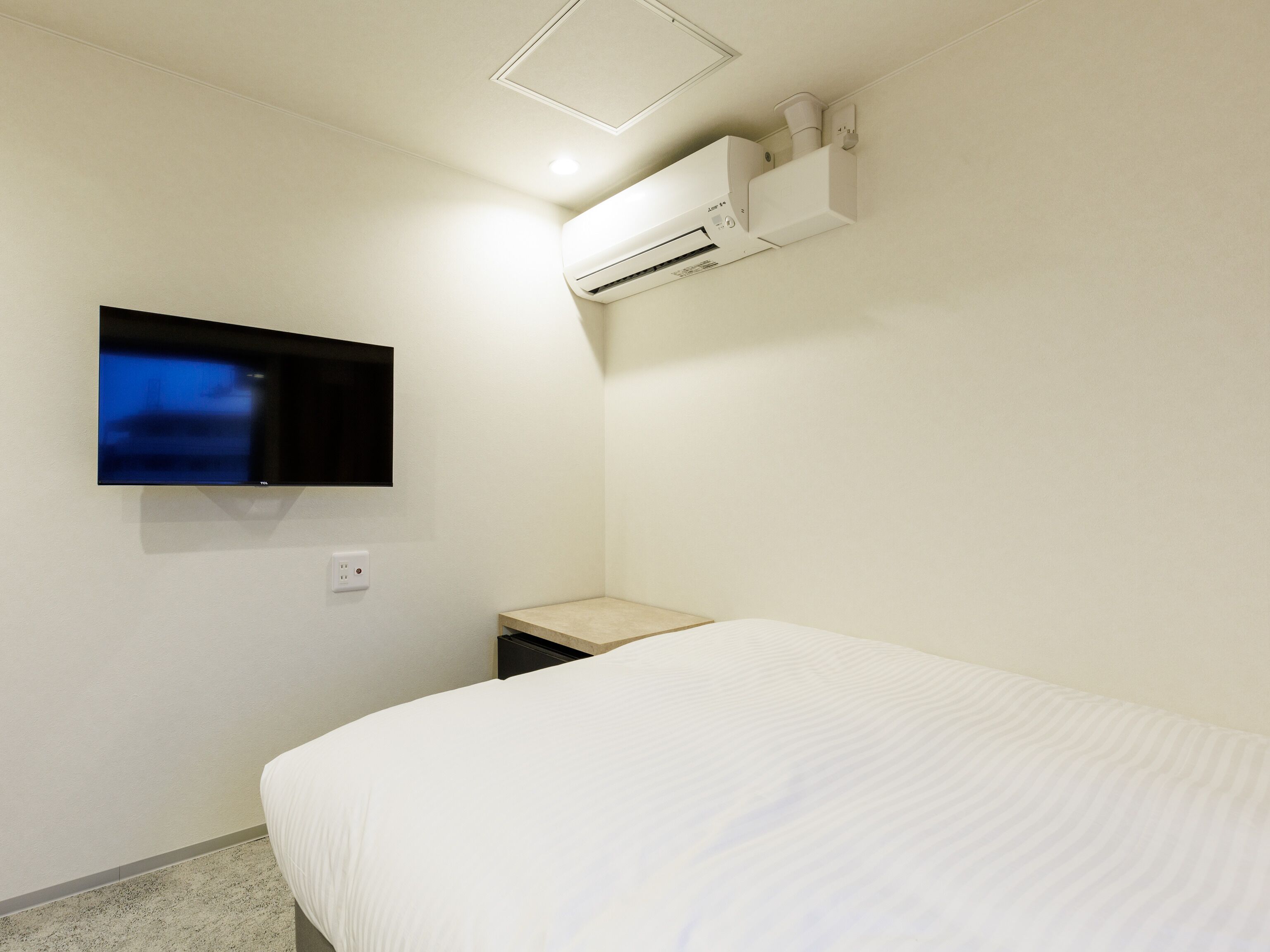 A hotel room with a bed, a mounted TV, and an air conditioning unit.