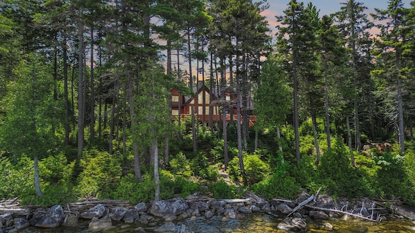 A lodge surrounded by dense forest, with a clear stream in the foreground. - Evergreen Point Lodge (Panoramic Views of Moosehead Lake!) (Greenville)