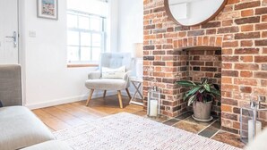 A cozy living room with a brick fireplace, a comfortable armchair, a large window, and a decorative mirror. - Beachcomber, Aldeburgh (Suffolk)