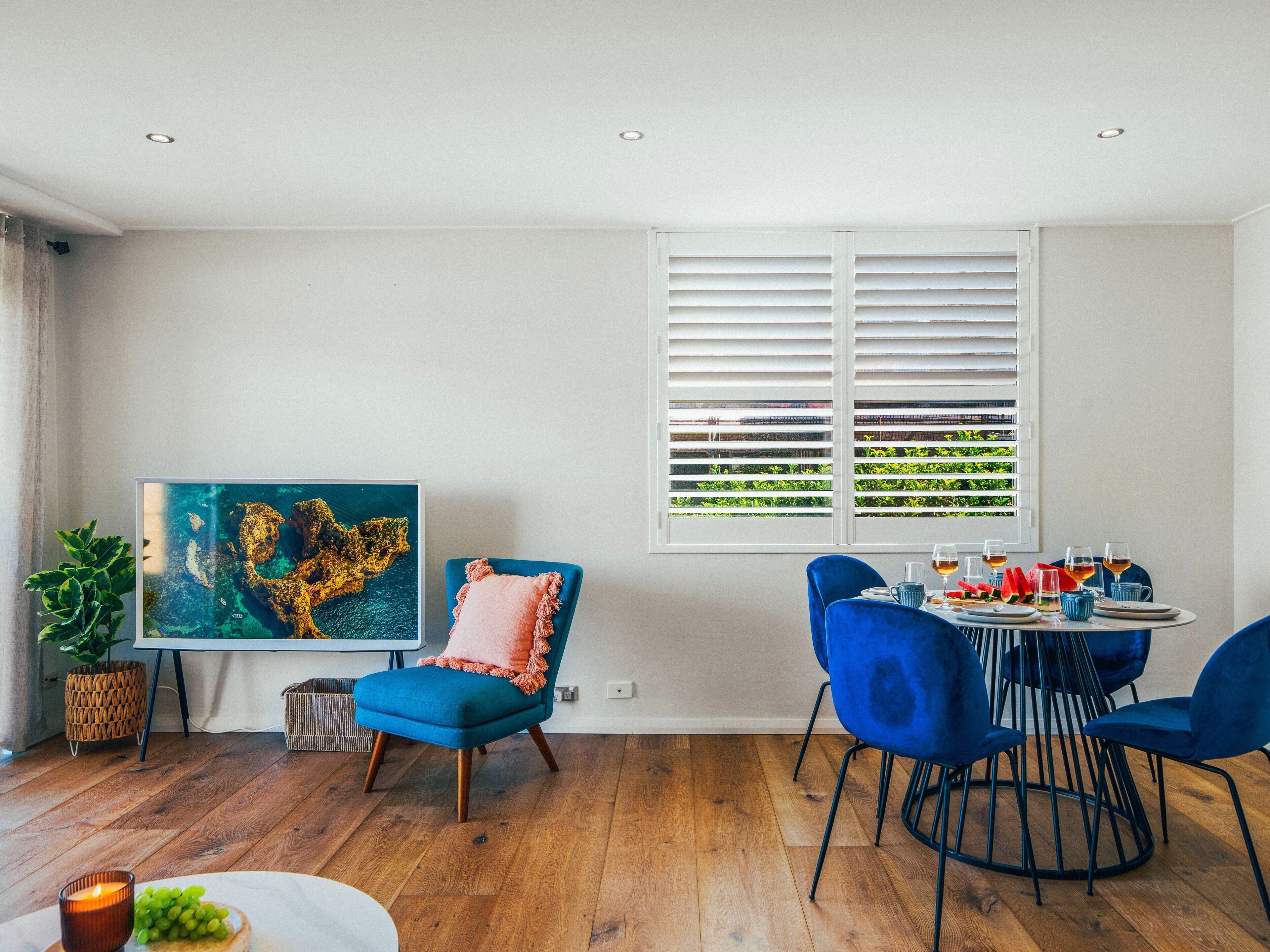A dining area with a table set for four, blue chairs, a large abstract painting, and a window with blinds.