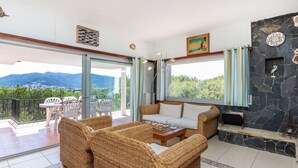 En rummelig opholdsstue med fletningsmøbler, en stenspir og en balkon med udsigt til et bjerglandskab.