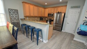 A kitchen with wooden cabinets, a stainless steel refrigerator, and a bar area with stools. - The Blue Pearl - Walk to the Beach in Garden City (Garden City Beach)