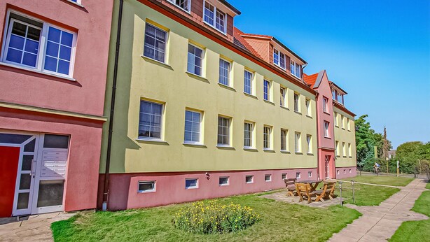 A multi-story building with a red and yellow facade, a small garden with yellow flowers, and a paved walkway.