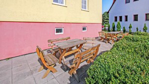 An outdoor seating area with wooden tables and benches, a pink wall, and a hedge.