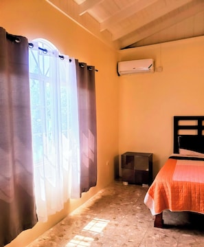A bedroom with a bed, a small dark-colored cabinet, a window with curtains, and an air conditioning unit on the wall. - Comfy Apartments (Montego Bay)