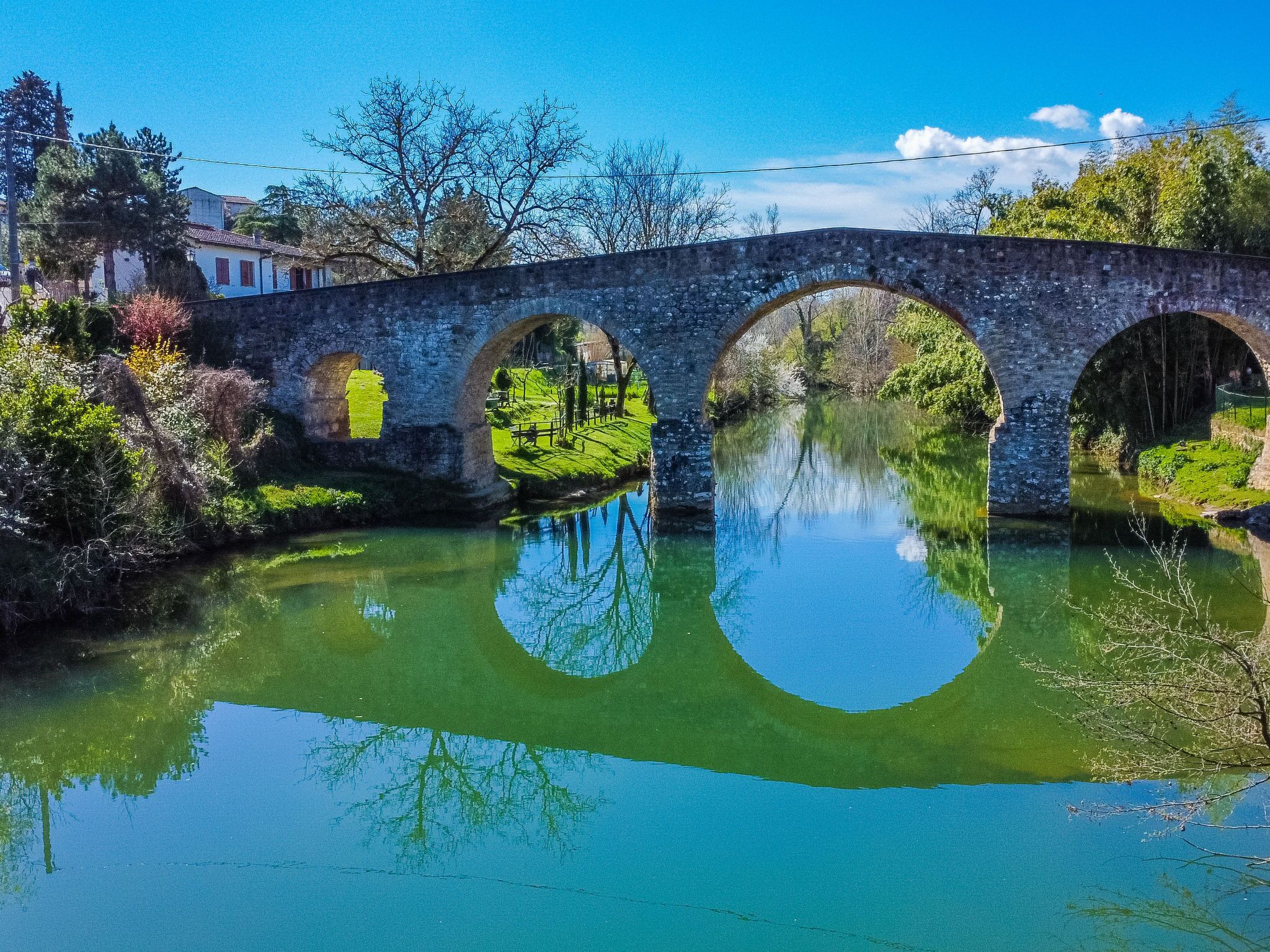 Un pont de pierre à trois arches enjambant une rivière paisible, entouré de verdure et d’arbres.