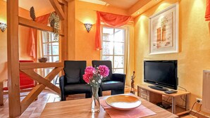 Un salon chaleureux avec une table en bois, deux chaises noires, un vase rempli de fleurs roses, un téléviseur et un tableau encadré accroché au mur.