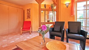 Une pièce avec un plancher en bois, deux chaises noires, une chaise rouge, une table en bois sur laquelle se trouvent un bol et des fleurs, et une fenêtre avec des rideaux.