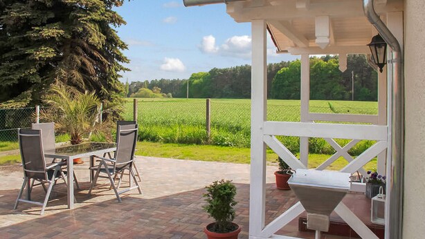 Een terras buiten met een tafel en stoelen, een potplant en een witte constructie met een raam.