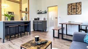 A modern living room with a kitchen area, wooden flooring, a coffee table with a remote control, a grey sofa, and a decorative clock on the wall. - Ferienwohnung Blankenhagen (Blankenhagen)