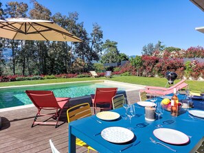 A poolside dining area with a table set for two, surrounded by red chairs, under a large umbrella.
