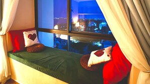 A cozy room with a window seat featuring a green cushion, red and white pillows, and a view of a cityscape at night.