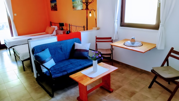 Una habitación con un sofá azul, una mesita de madera, una silla, una mesa de comedor con un pastel y una ventana con cortinas.