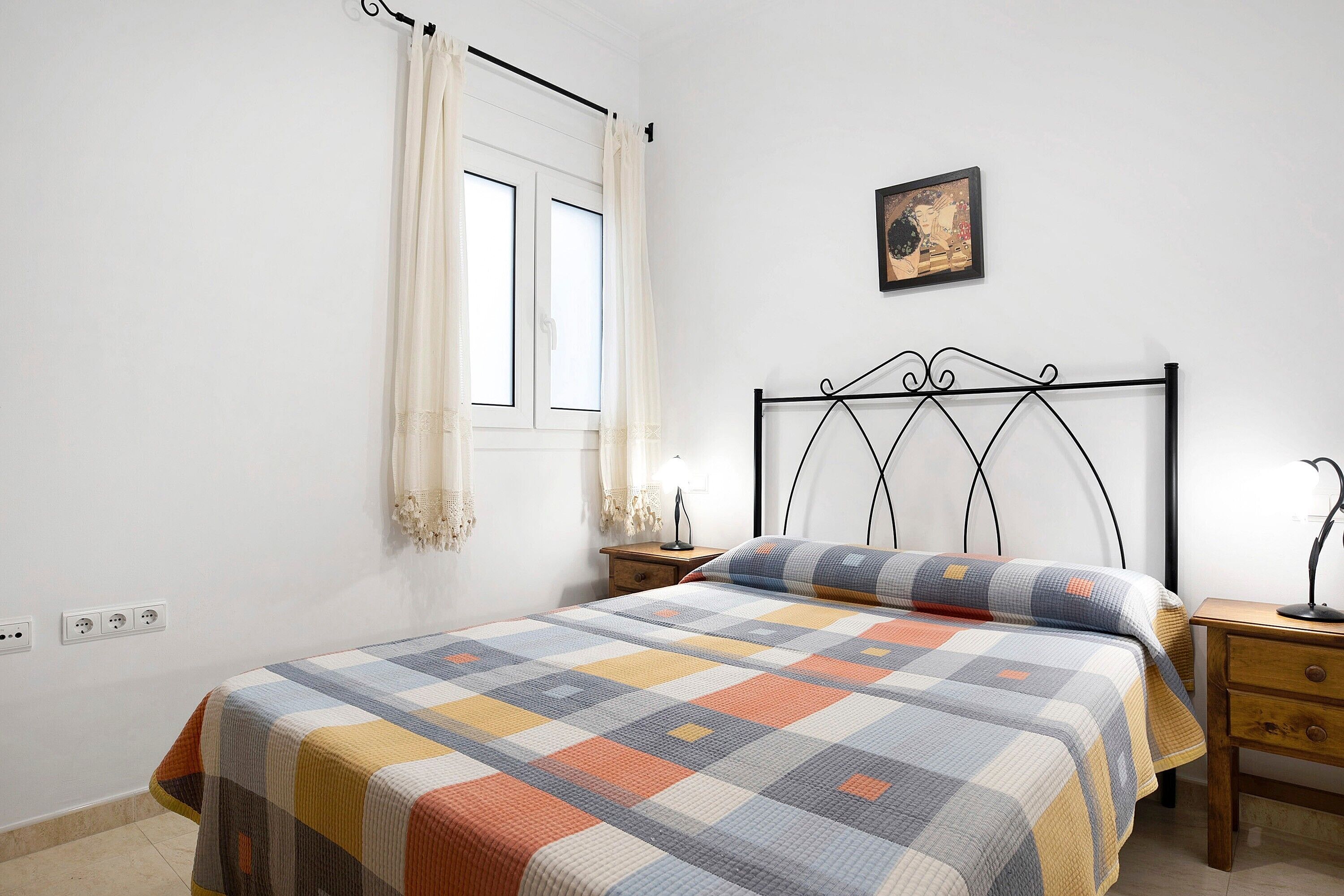 A bedroom with a bed, a nightstand, a lamp, a window with curtains, and a wall-mounted picture.