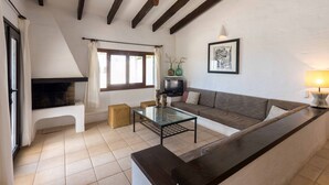 Ein Wohnzimmer mit einer Couch, einem Glastisch und einem Fernseher. Es gibt einen Kamin, einen Deckenventilator und ein Fenster mit Vorhängen.