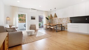 A modern living room with a sofa, dining table, and chairs, a kitchen with white cabinets, and a view of greenery outside. - Apartment 'Casa Aga Seaside Chic' with Balcony, Wi-Fi and Air Conditioning (Ericeira)