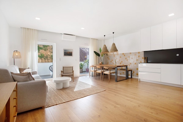 A modern living room with a sofa, dining table, and chairs, a kitchen with white cabinets, and a view of greenery outside.