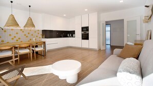 A modern kitchen with white cabinets, a dining area with a wooden table and chairs, and a living area with a sofa and cushions. - Apartment 'Casa Aga Seaside Chic' with Balcony, Wi-Fi and Air Conditioning (Ericeira)
