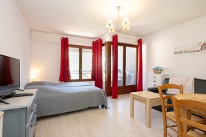 A bedroom with a bed, a desk, a chair, a TV, and a window with curtains.