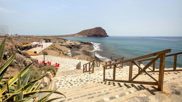 Une plage avec un escalier descendant vers le rivage, un promontoire rocheux à l’horizon et un ciel dégagé.