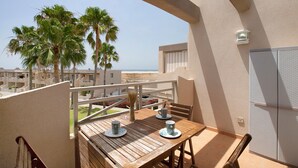 Un balcon avec une table et des chaises en bois, une vue sur les palmiers et la mer, et un bâtiment doté d’un mur à texture et d’un luminaire.