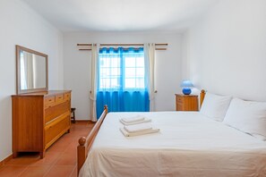 Un dormitorio con cama, cómoda, mesita de noche, lámpara y ventana con cortinas.