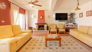 A living room with a sofa, armchairs, a coffee table, a fireplace, a television, and a ceiling fan. - Villa 'Kethellen' with Sea View, Private Terrace and Wi-Fi (Porto Covo)