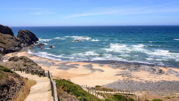 Une plage côtière avec un escalier en bois descendant jusqu’au rivage, des affleurements rocheux et l’océan.