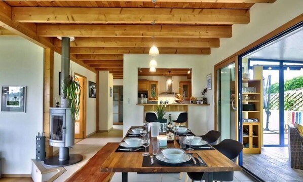A dining area with a wooden table set for four, a kitchen in the background, and a glass door leading to an outdoor area. - Spacious House – Sea View, 2 Minutes from GR34 and Beaches (Plouarzel)