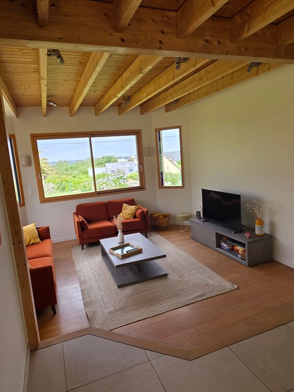 Ein Wohnzimmer mit Holzboden, einer Couch, einem Couchtisch, einem Fernseher und großen Fenstern.