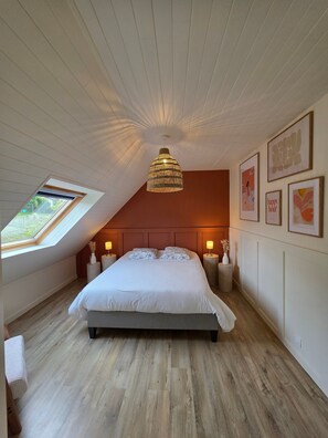 Ein Schlafzimmer mit schrägen Wänden, einem Oberlicht, einem Bett mit zwei Kissen, Nachttischlampen und gerahmten Kunstwerken an den Wänden.