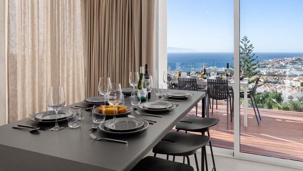 Une table à manger dressée pour un repas, avec vue sur la ville, de grandes fenêtres et un balcon.